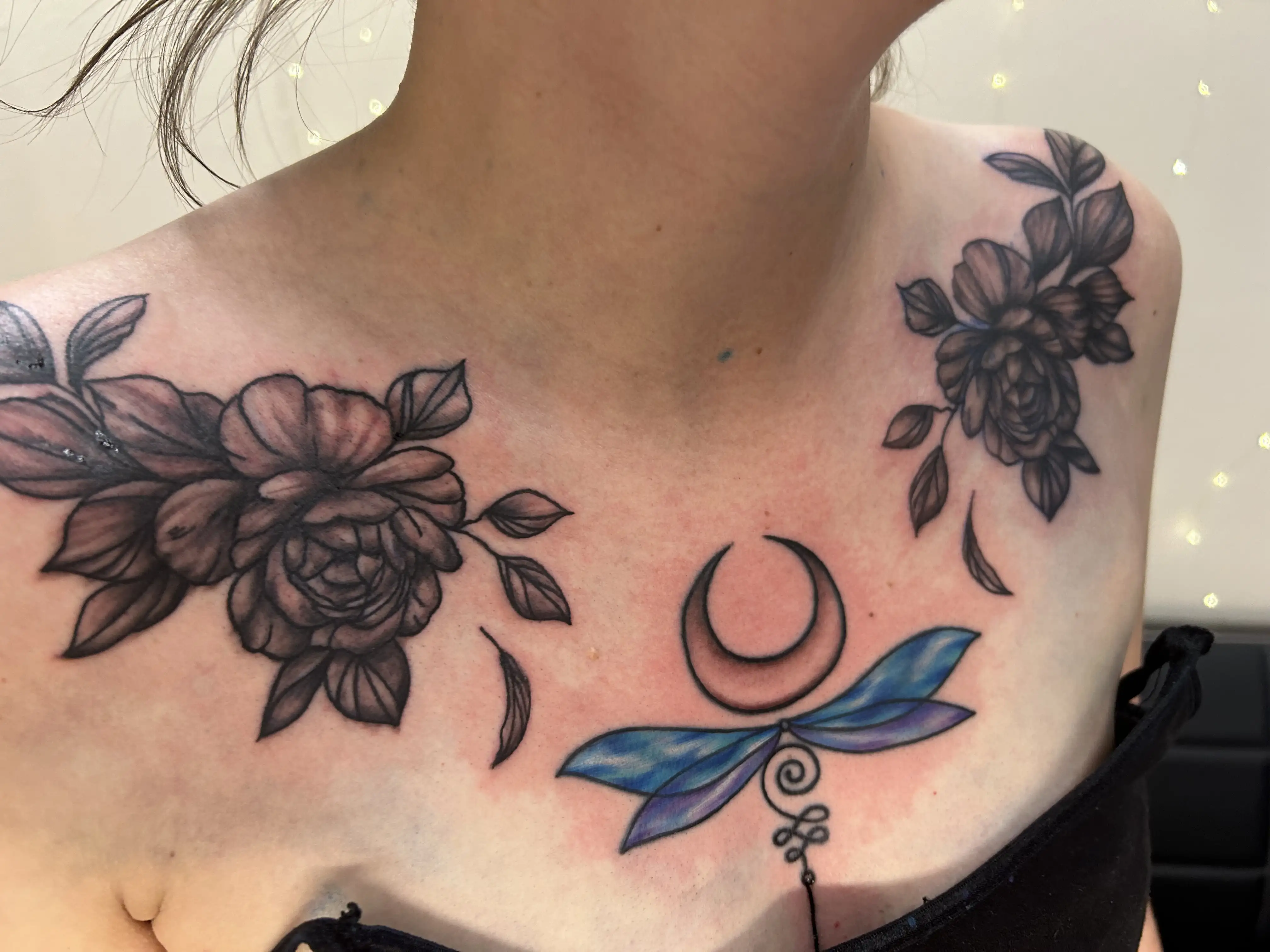 BL-ink Tattoo Colorado Springs chest set: black-grey peony clusters on both collarbones framing a central piece of a blue-to-violet dragonfly beneath a crescent moon.